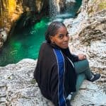 A detailed view of Nma Okafor sitting on rocks beside a scenic waterfall in Slovenia, showcasing her enjoying nature and outdoor adventure in a picturesque European landscape.