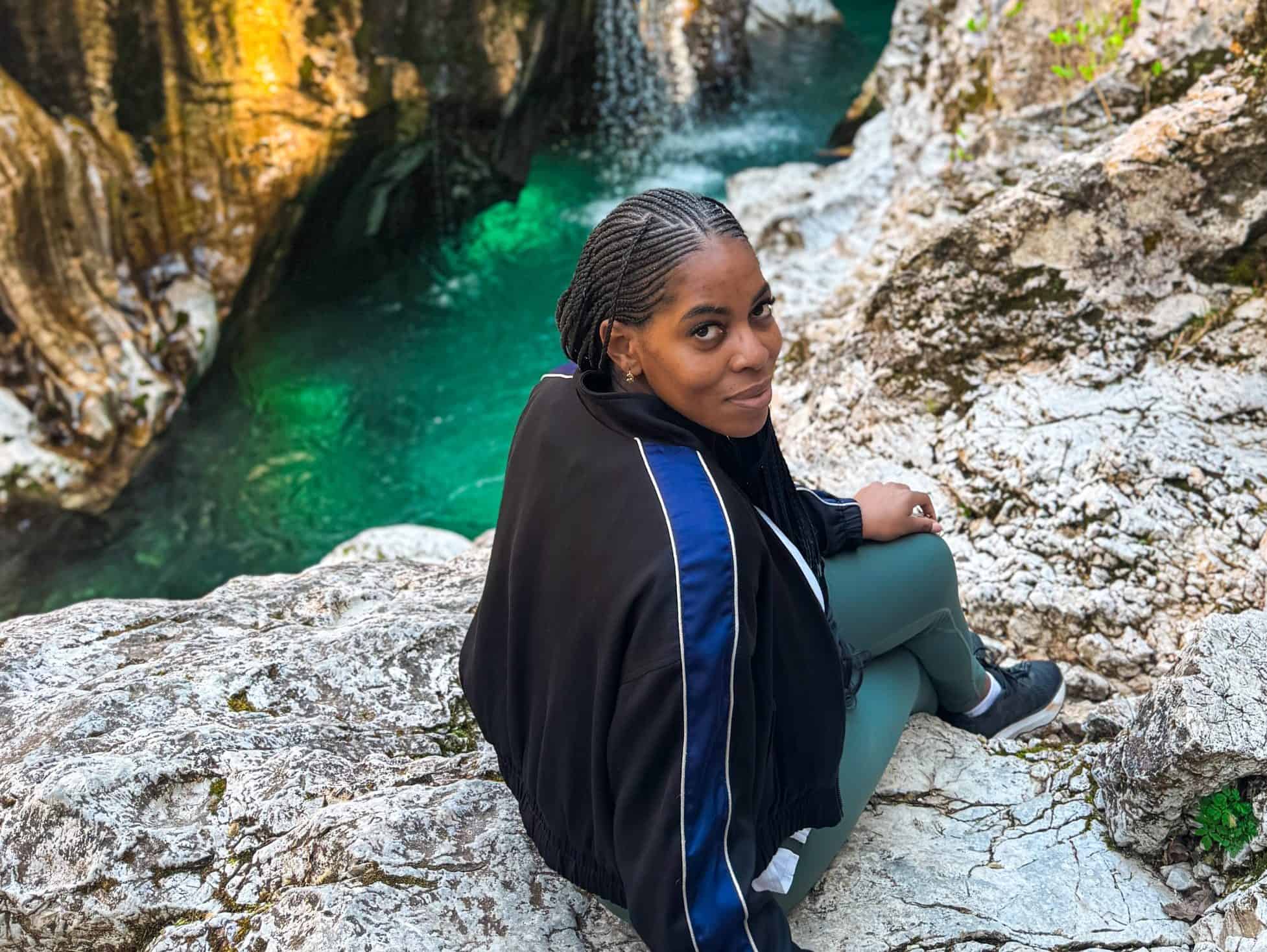 A detailed view of Nma Okafor sitting on rocks beside a scenic waterfall in Slovenia, showcasing her enjoying nature and outdoor adventure in a picturesque European landscape.