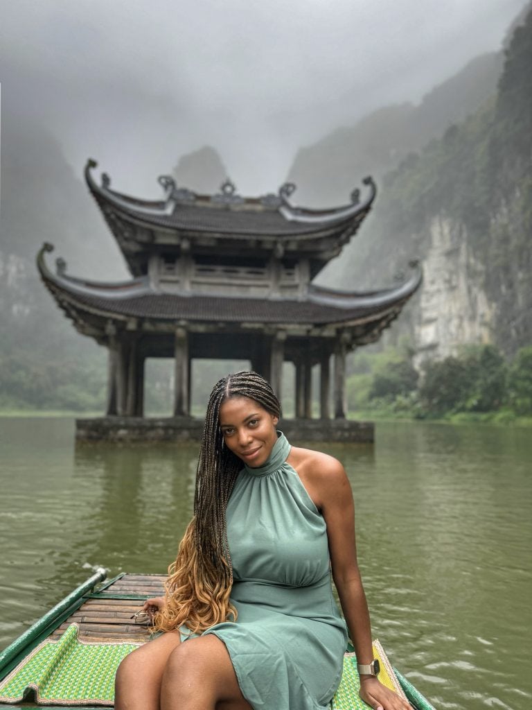 Serene woman with braided hair enjoying a boat ride on a river in Vietnam, with a traditional pagoda-style structure and misty mountains in the background, capturing beauty and tranquility.