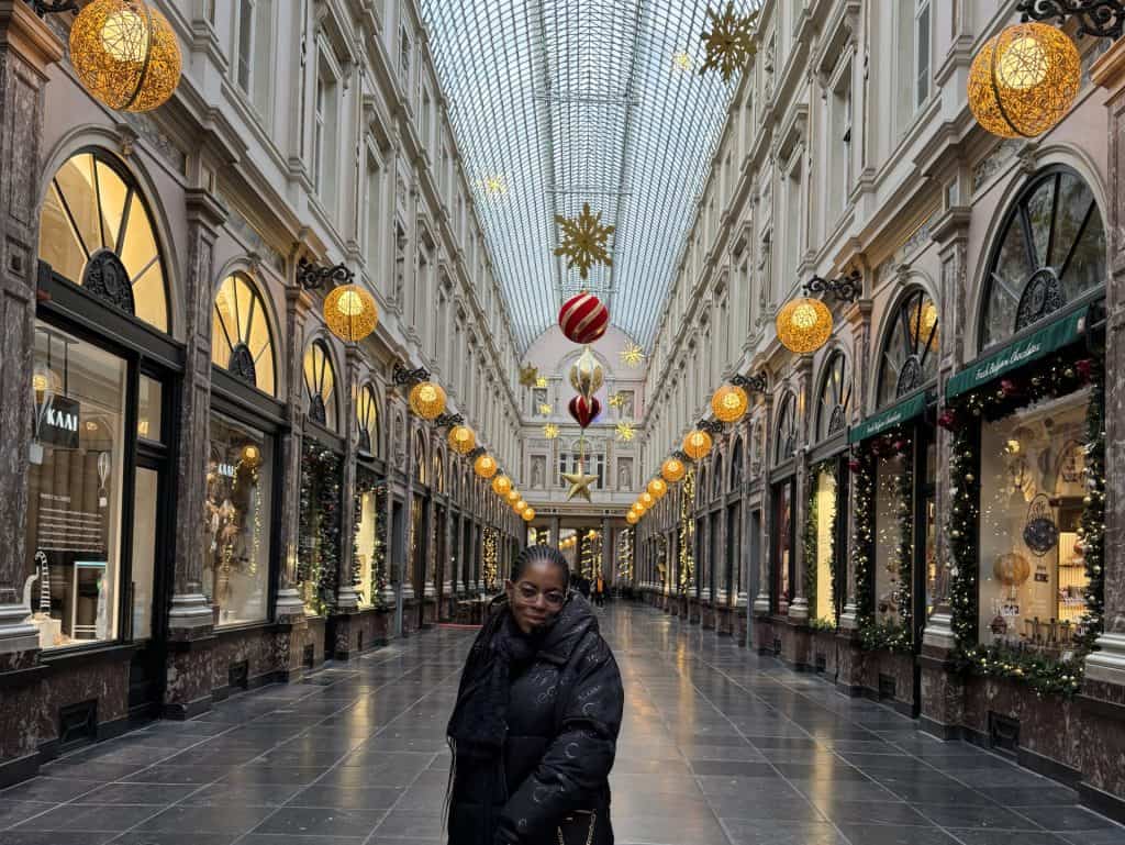 Elegant woman standing inside a beautifully decorated shopping arcade during Christmas season, with festive ornaments and warm lighting, showcasing holiday shopping and fashion.