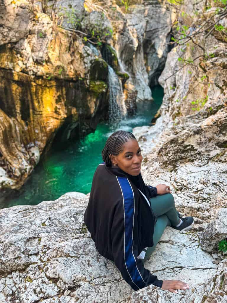 Vibrant outdoor portrait of Nma Okafor sitting on rocky terrain near a waterfall, showcasing her connection to nature, adventure, and exploration, with stunning green water and rugged cliffs in the background.