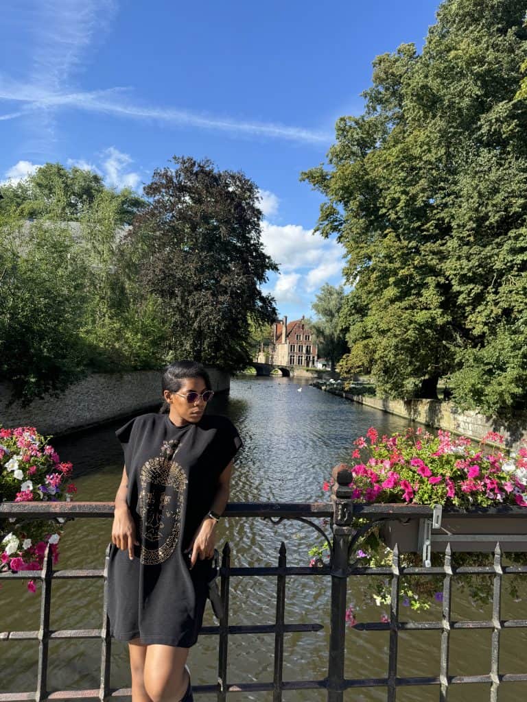 A woman in a black graphic dress and sunglasses standing by a river with lush trees and historic buildings in the background, enjoying a sunny day in a scenic European city - Brugges.