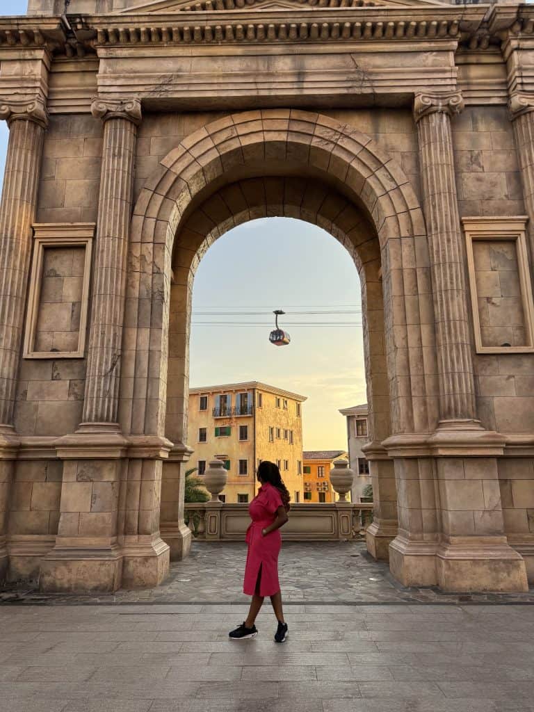 Elegant woman in pink dress standing under ancient architectural arch at sunset, showcasing African-inspired fashion and cultural heritage, with a cityscape in the background.