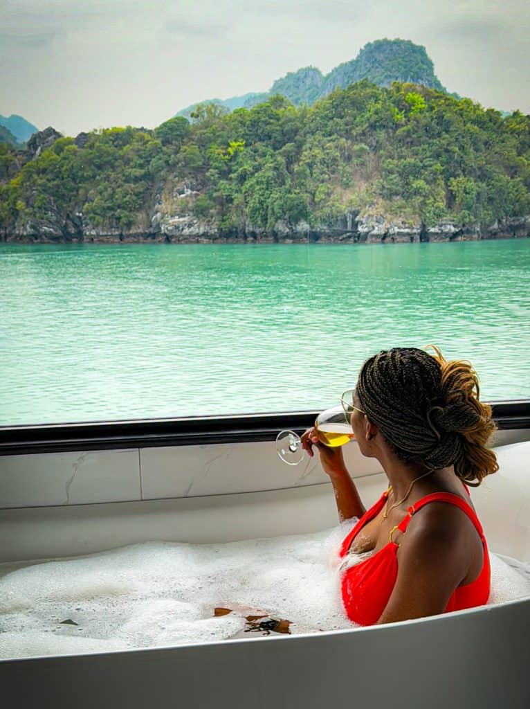 Relaxing woman enjoying a glass of wine in a bathtub overlooking a serene lake and lush green mountains, emphasizing tranquility, luxury, and nature connection.