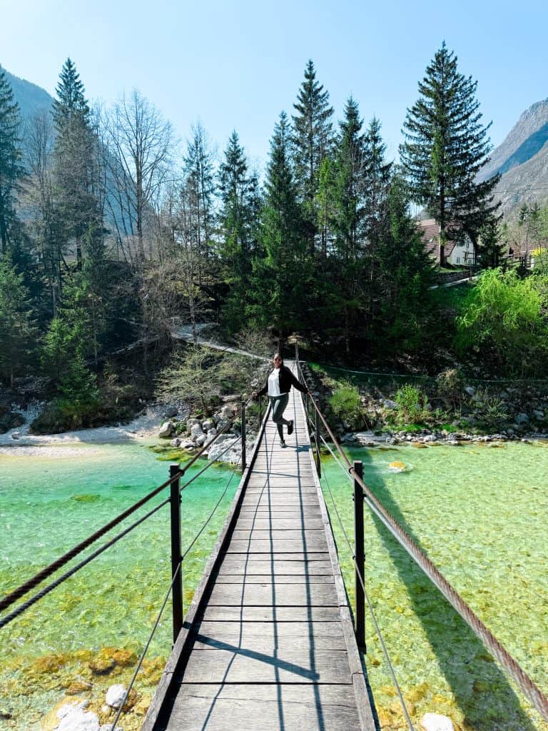 Suspension bridge over a turquoise river surrounded by lush green trees and mountains in the background, with a woman standing confidently on the bridge, enjoying the scenic outdoor landscape.