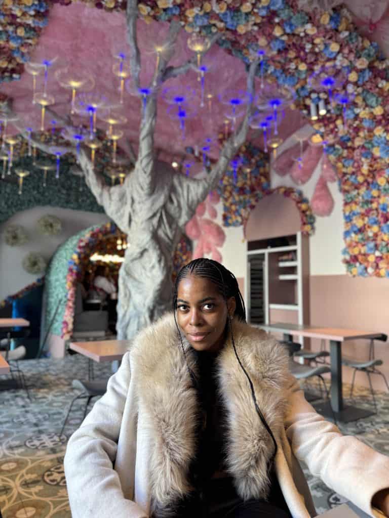 Elegant woman with braided hair wearing a faux fur coat in a creatively designed floral-themed restaurant, vibrant ceiling decor, and artistic interior accents, embodying style, confidence, and modern fashion.