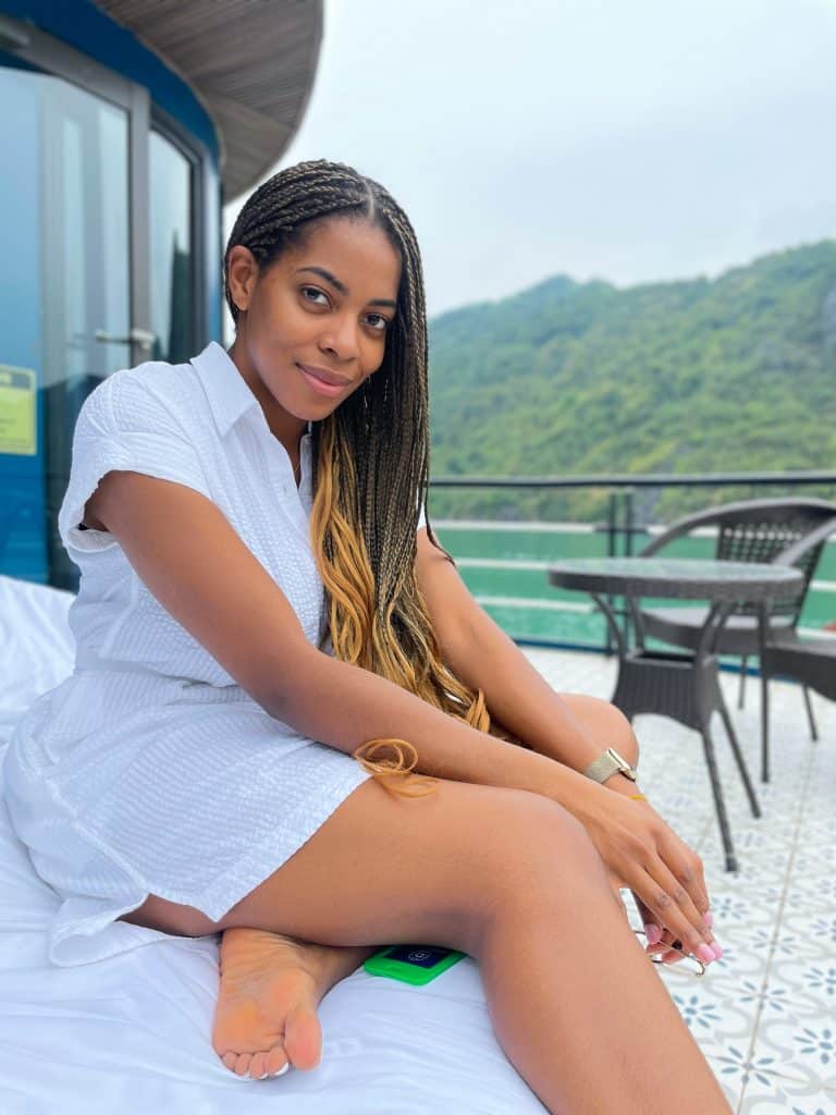 Relaxed African woman with braids sitting on a bed overlooking a scenic water and mountain view, wearing a white dress, enjoying a peaceful moment outdoors.