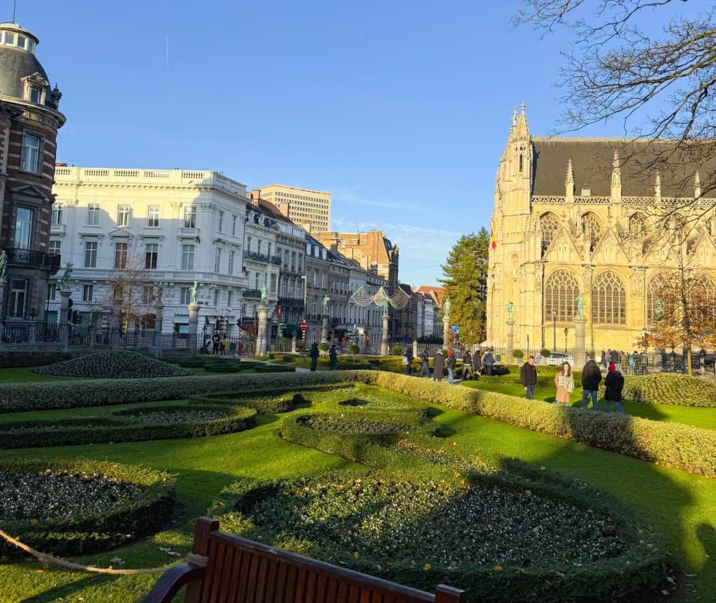 Vibrant cityscape featuring historic buildings and a beautiful Gothic church with a lush, well-maintained garden in the foreground, showcasing the scenic beauty and architecture of Brussels, Belgium.