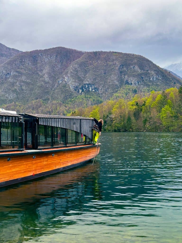This image features a peaceful mountain lake with a wooden boat and a person enjoying outdoor adventure amidst vibrant green foliage and towering mountains under a overcast sky, highlighting nature, travel, and scenic landscapes.