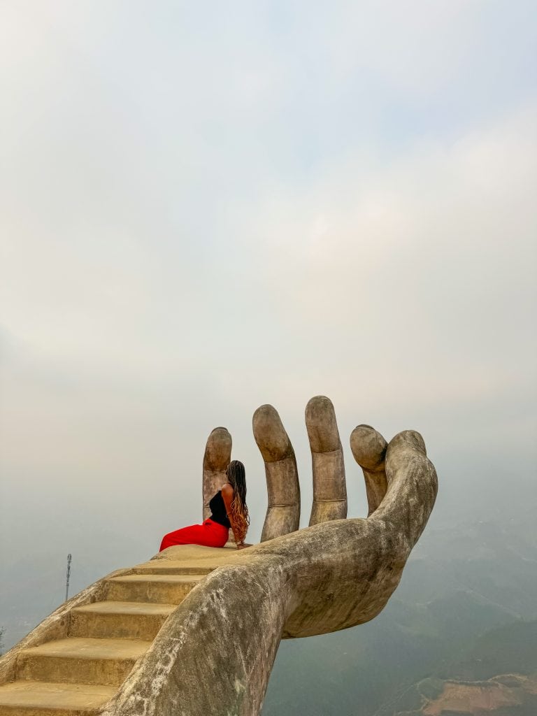 Bold sculpture of a hand with the woman in stylish clothing sitting on the stairs, emphasizing artistic expression and cultural heritage.
