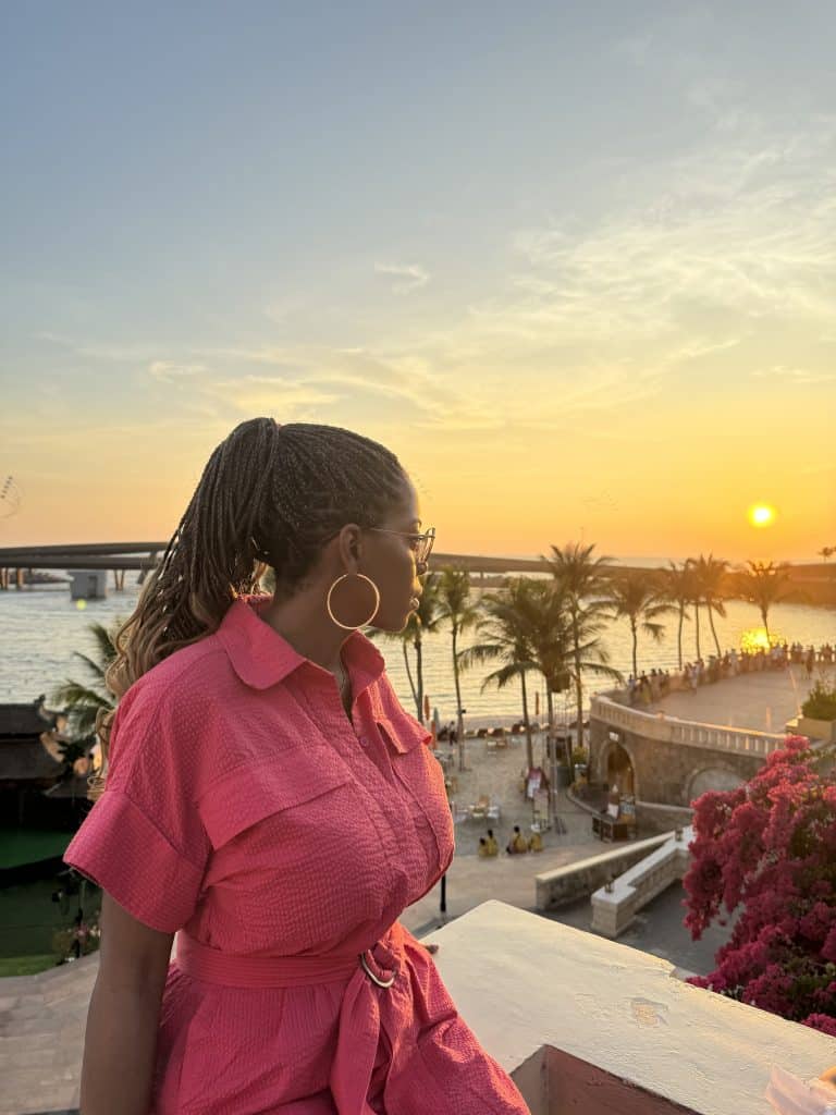 Elegant woman with braided hair and large hoop earrings enjoying a sunset view at a tropical waterfront resort, showcasing stylish fashion and scenic beauty.