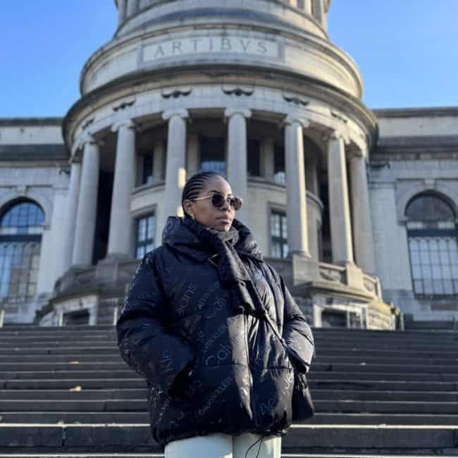 Elegant woman in designer jacket and sunglasses standing on steps outside historic building in London, showcasing Nma Okafor’s fashion influence and urban style in a scenic architectural setting.