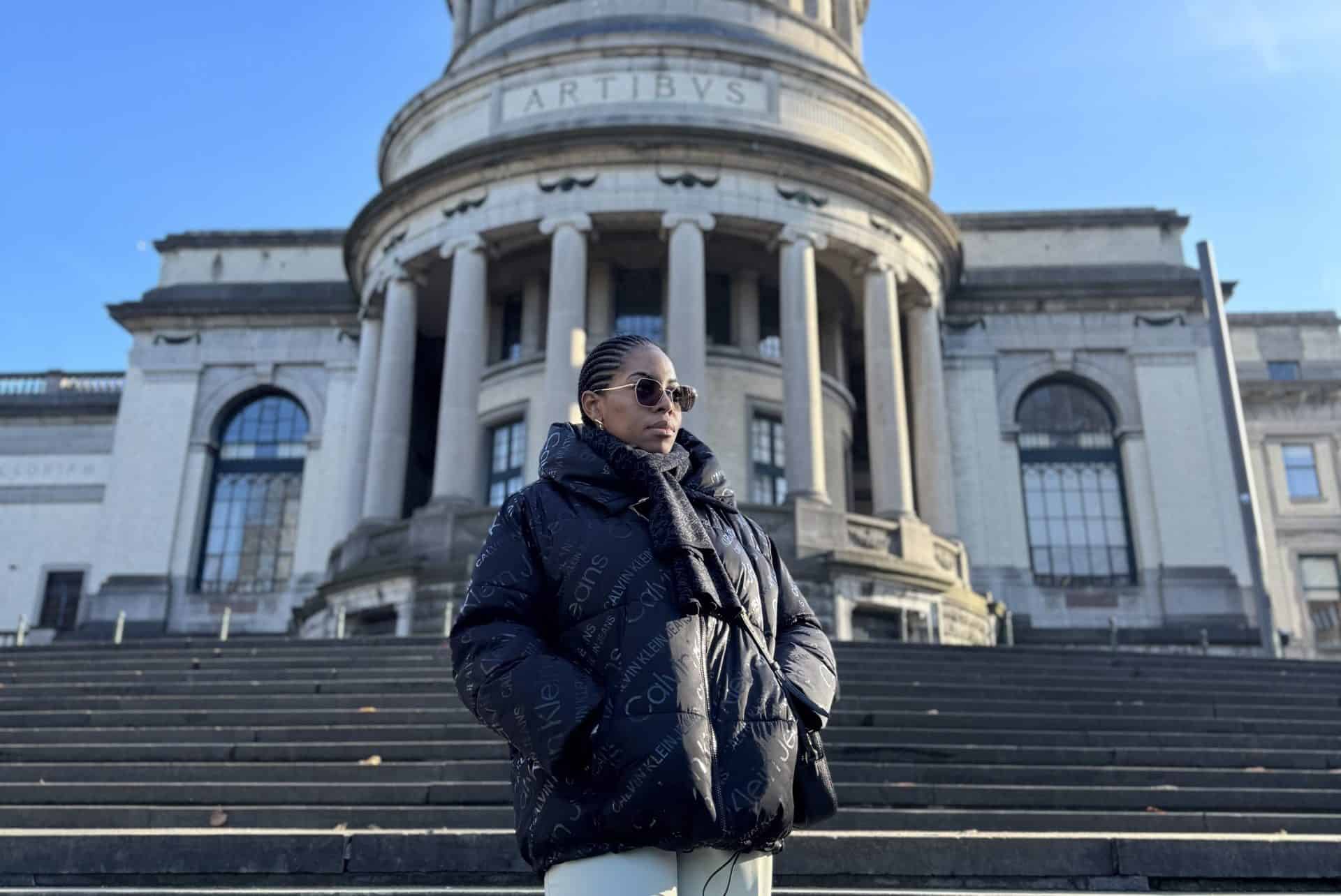 Elegant woman in designer jacket and sunglasses standing on steps outside historic building in London, showcasing Nma Okafor’s fashion influence and urban style in a scenic architectural setting.