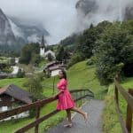 A woman in a pink dress walking along a mountain trail in Switzerland, surrounded by lush greenery, traditional houses, and misty mountain peaks in the background.