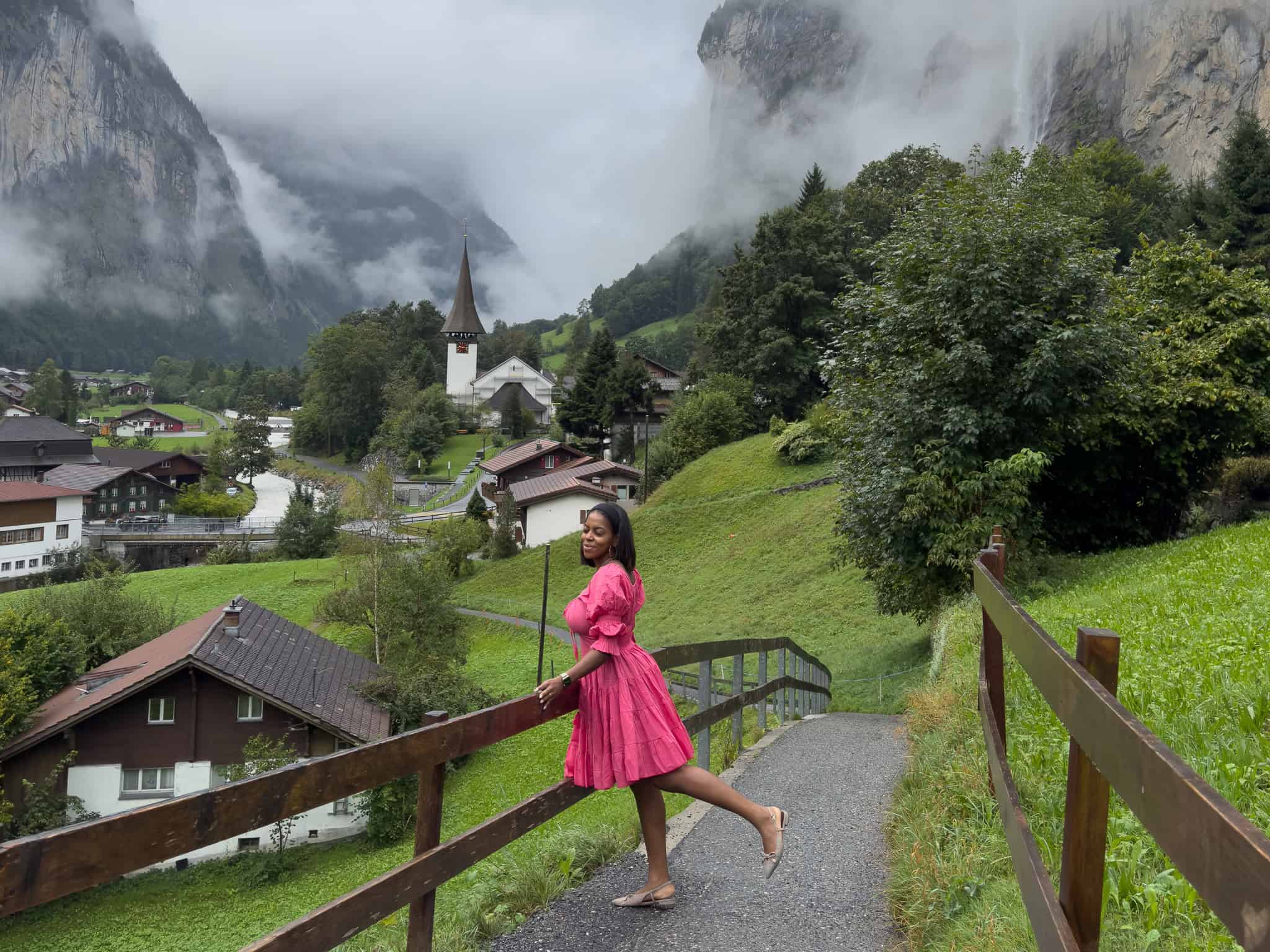 A woman in a pink dress walking along a mountain trail in Switzerland, surrounded by lush greenery, traditional houses, and misty mountain peaks in the background.