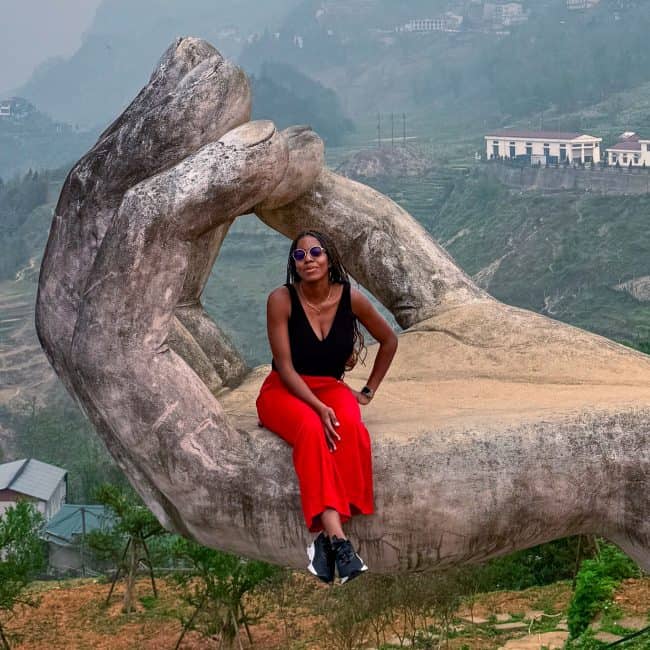A woman sitting on a large, artistic hand sculpture overlooking lush green terraced fields and mountains in Vietnam, showcasing cultural artistry and scenic beauty.