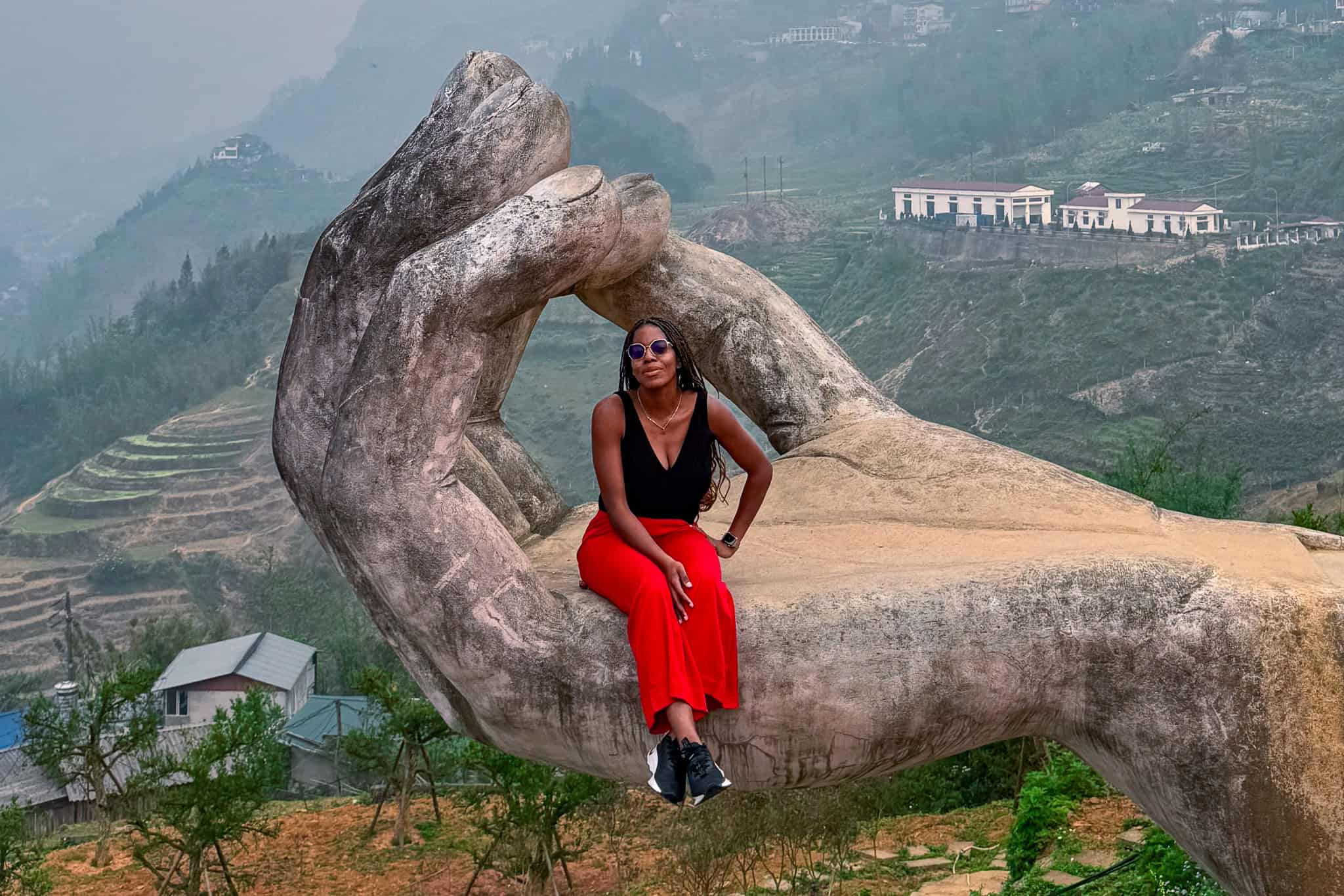 A woman sitting on a large, artistic hand sculpture overlooking lush green terraced fields and mountains in Vietnam, showcasing cultural artistry and scenic beauty.
