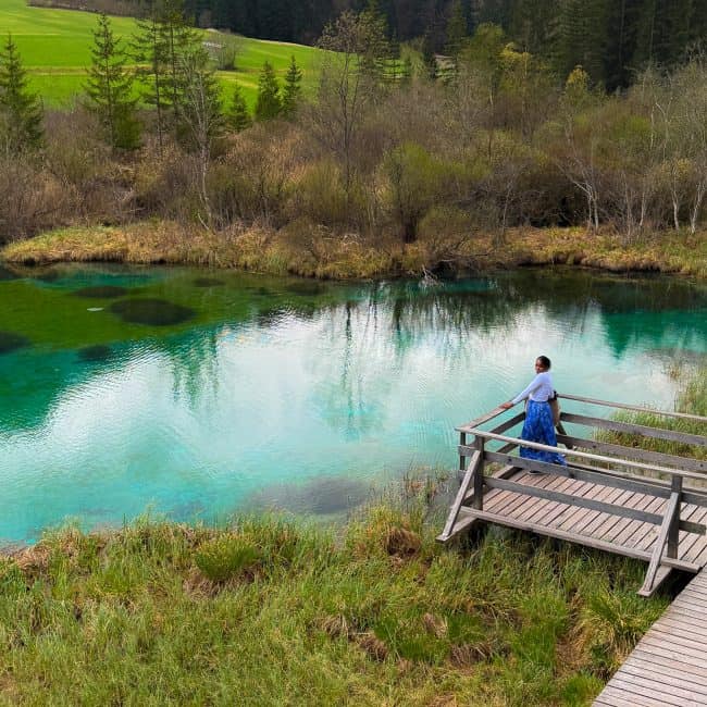 Scenic view of a turquoise lake in Slovenia with a person walking on a wooden pathway surrounded by lush greenery and trees.