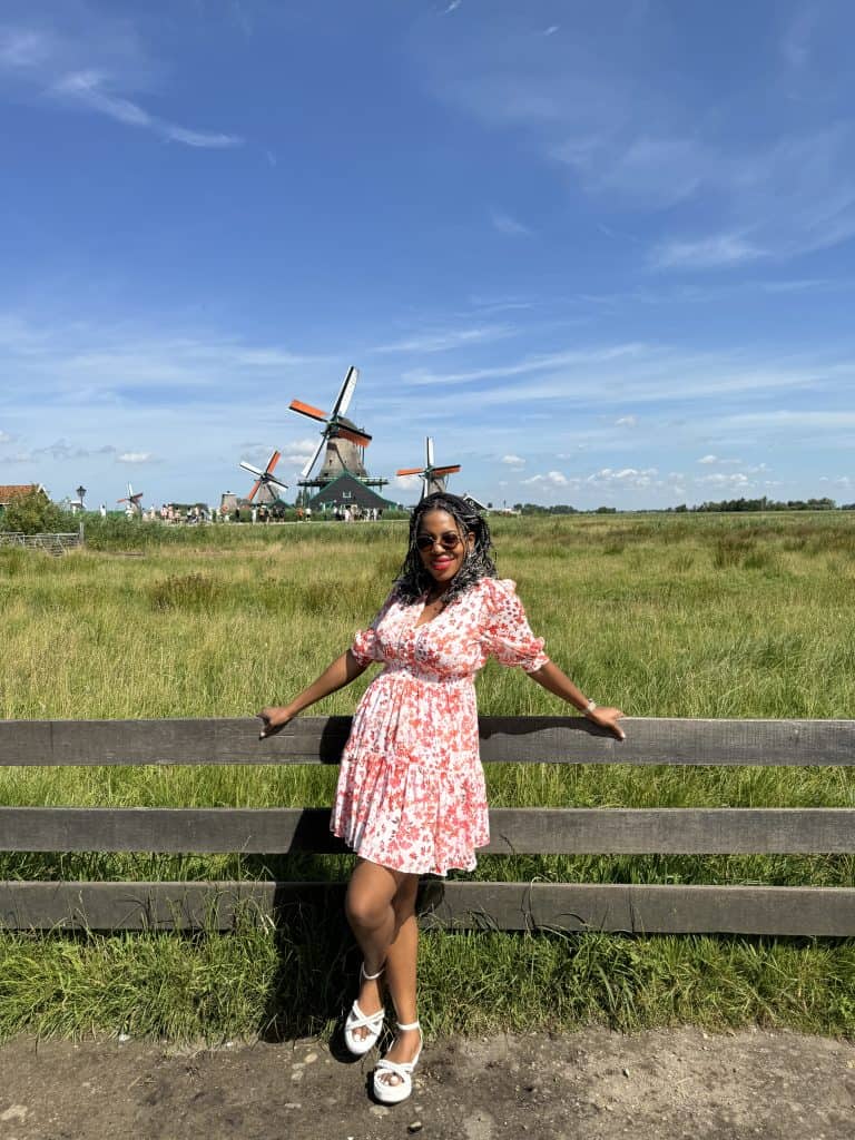 Woman in a floral dress standing in front of a wooden fence with windmills in the background under a blue sky, showcasing Dutch countryside scenery.