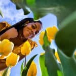 A woman smiling and enjoying a sunny day among vibrant yellow tulips in a lush garden setting.