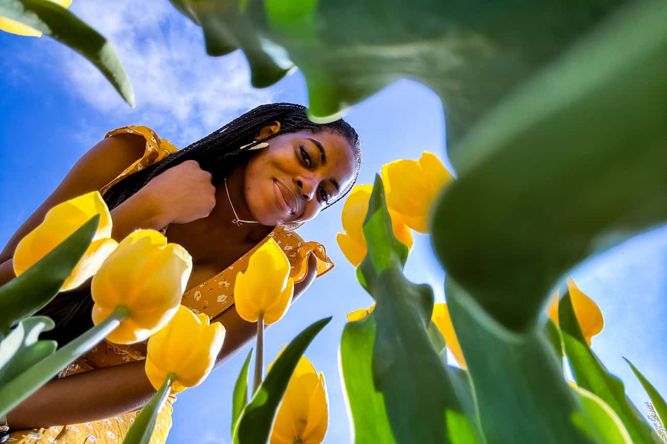 A woman smiling and enjoying a sunny day among vibrant yellow tulips in a lush garden setting.
