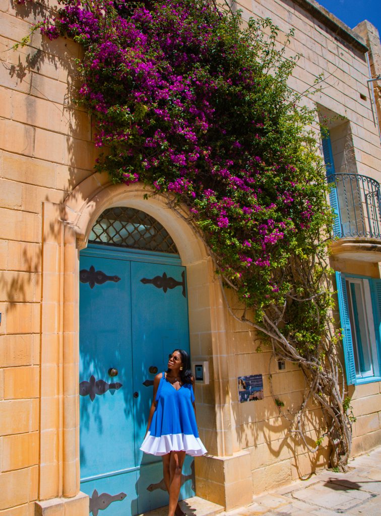 Nma Okafor standing in front of a vibrant blue door with a lush purple bougainvillea climbing the historic stone building. She wears a bright blue dress with white accents, showcasing elegance and sty.