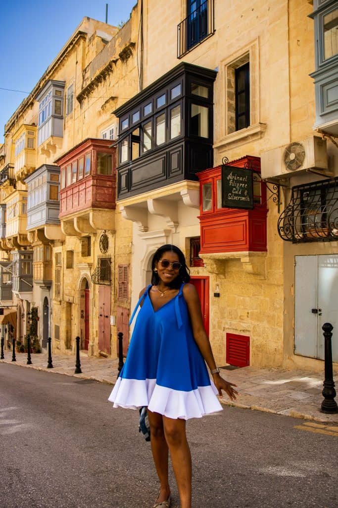 Nma Okafor standing on a vibrant Malta street with traditional balconies, wearing a blue and white dress, sunglasses, and smiling at the camera.