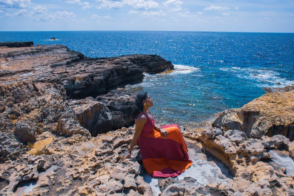 Nma Okafor relaxing on the rocky coast at Dwerja Bay, gazing at the ocean, with clear blue skies and waves crashing against the rocks, capturing a peaceful seaside moment.