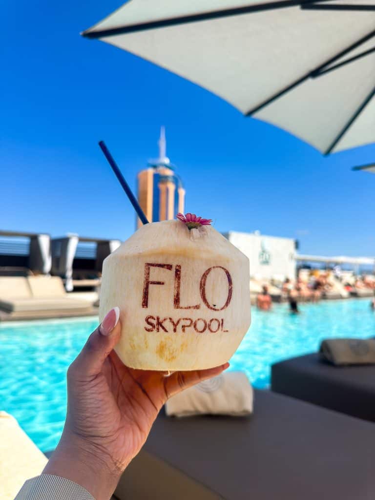 Refreshing coconut beverage with a straw, featuring the words "FLO SKYPool" carved on the shell, set against a vibrant poolside scene with a city skyline in the background.