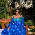 Nma Okafor sitting on a wooden bench in a lush garden, wearing sunglasses and a bright blue dress with floral patterns, enjoying a sunny day surrounded by greenery and colorful flowers.