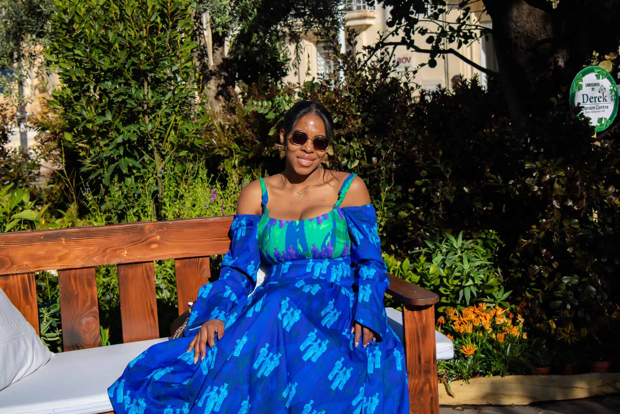 Nma Okafor sitting on a wooden bench in a lush garden, wearing sunglasses and a bright blue dress with floral patterns, enjoying a sunny day surrounded by greenery and colorful flowers.