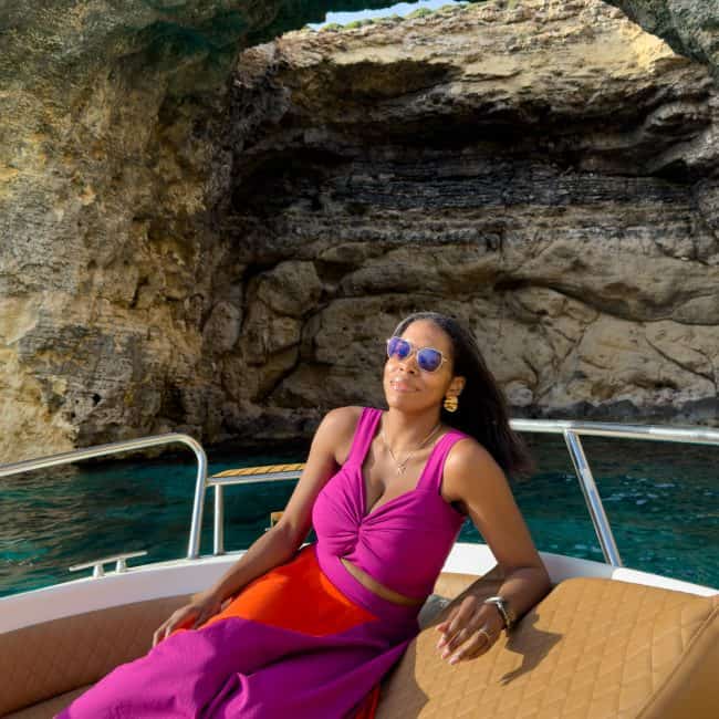 Nma Okafor relaxing on a boat near a rocky coastal cave, wearing stylish sunglasses and a vibrant purple and orange dress, showcasing a luxurious and adventurous lifestyle.