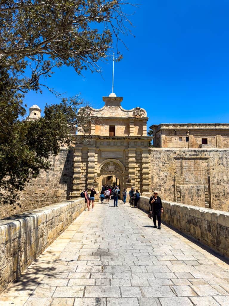 Historic fort entrance with visitors walking on stone pathway, showcasing colonial architecture and lush greenery under a clear blue sky.