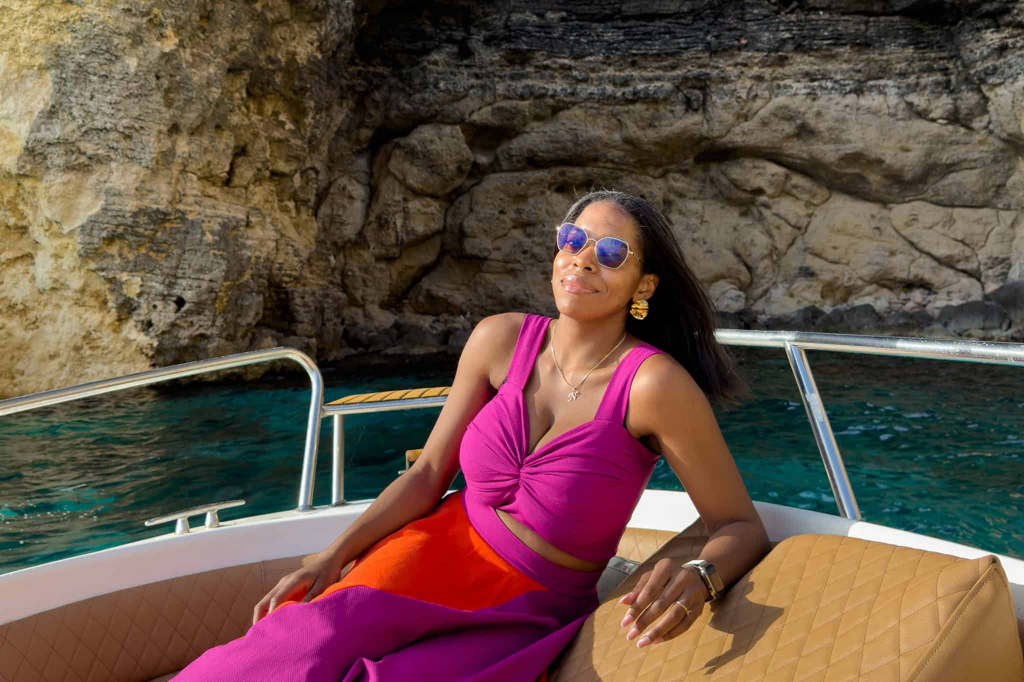 Nma Okafor relaxing on a boat in Malta, wearing sunglasses and a vibrant pink and orange dress.