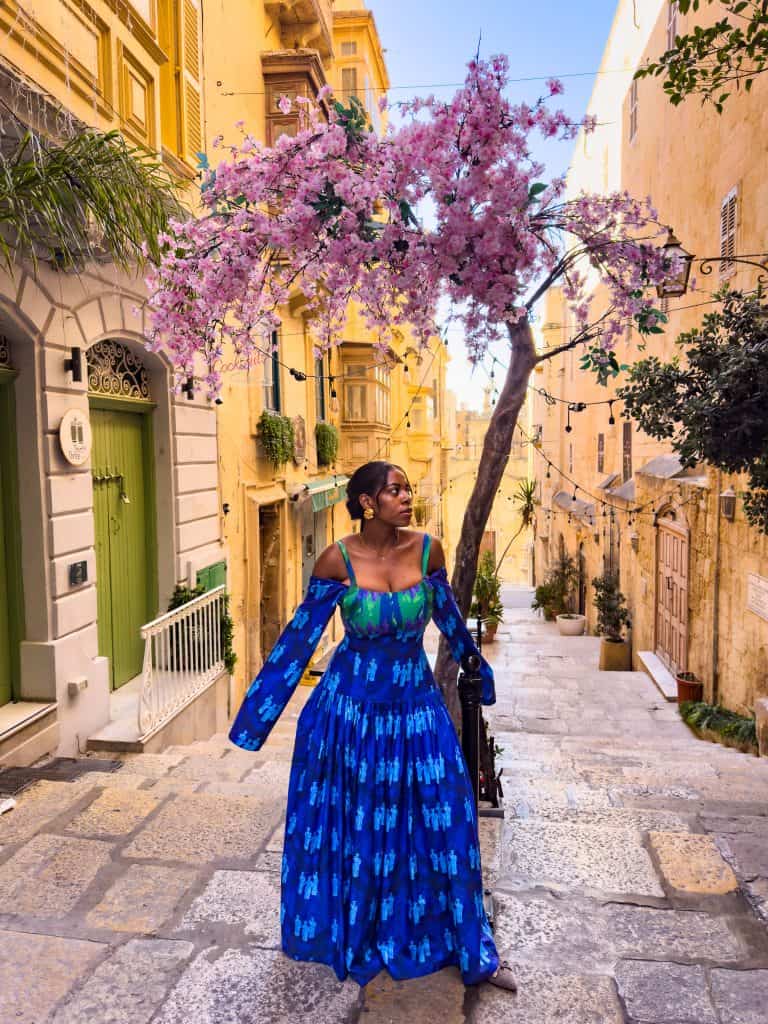 A stylish woman in a striking blue dress explores a picturesque street in Malta, showcasing the island's unique blend of historic architecture and lively street life.