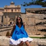 A woman in a vibrant blue and white dress sitting on a stone wall in front of historic fortress walls under a clear blue sky.