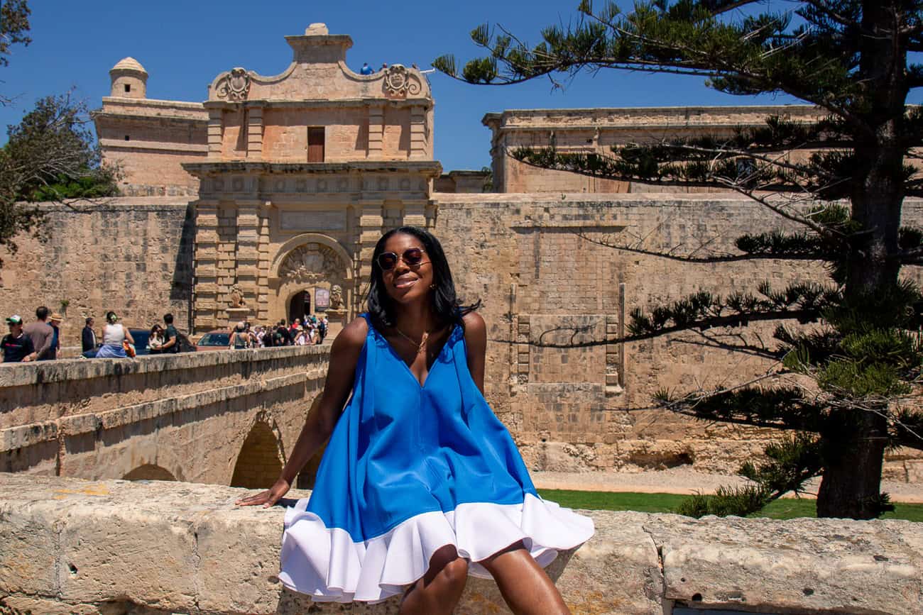 A woman in a vibrant blue and white dress sitting on a stone wall in front of historic fortress walls under a clear blue sky.