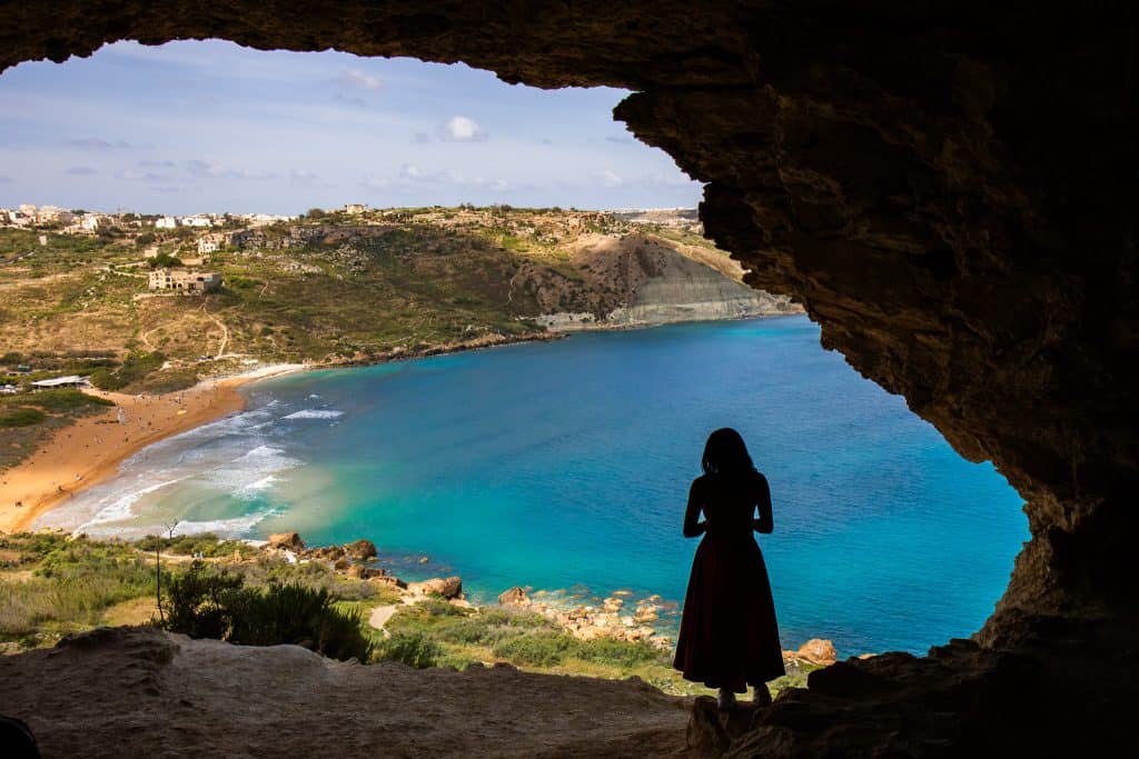 Woman standing at the edge of a cave, gazing at a stunning turquoise bay with sandy beaches and lush hills in the background.