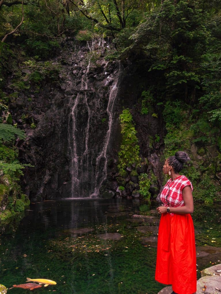 Nma Okafor standing near a waterfall in a lush green forest, wearing a red skirt and checkered top, enjoying nature in Japan.