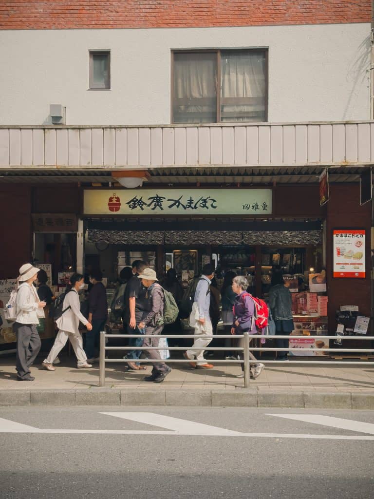 Crowd of people walking past a shop in Osaka, Japan, during daytime, with diverse attire and backpacks, under a building with Japanese signage.