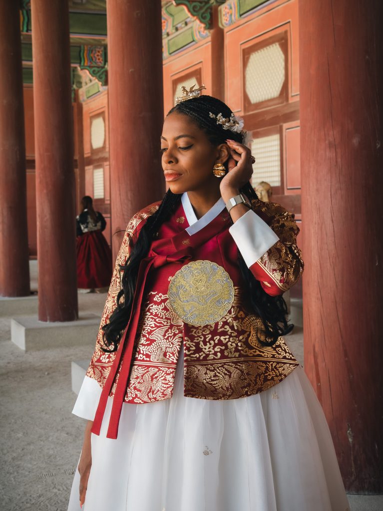 Woman wearing a vibrant traditional Korean hanbok with intricate embroidery, standing in a historic palace setting with colorful wooden architecture.