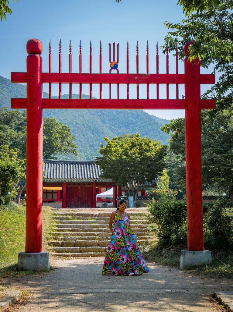A vibrant red traditional Korean gate with a woman in colorful hanbok standing beneath it, surrounded by lush greenery and mountains in the background.