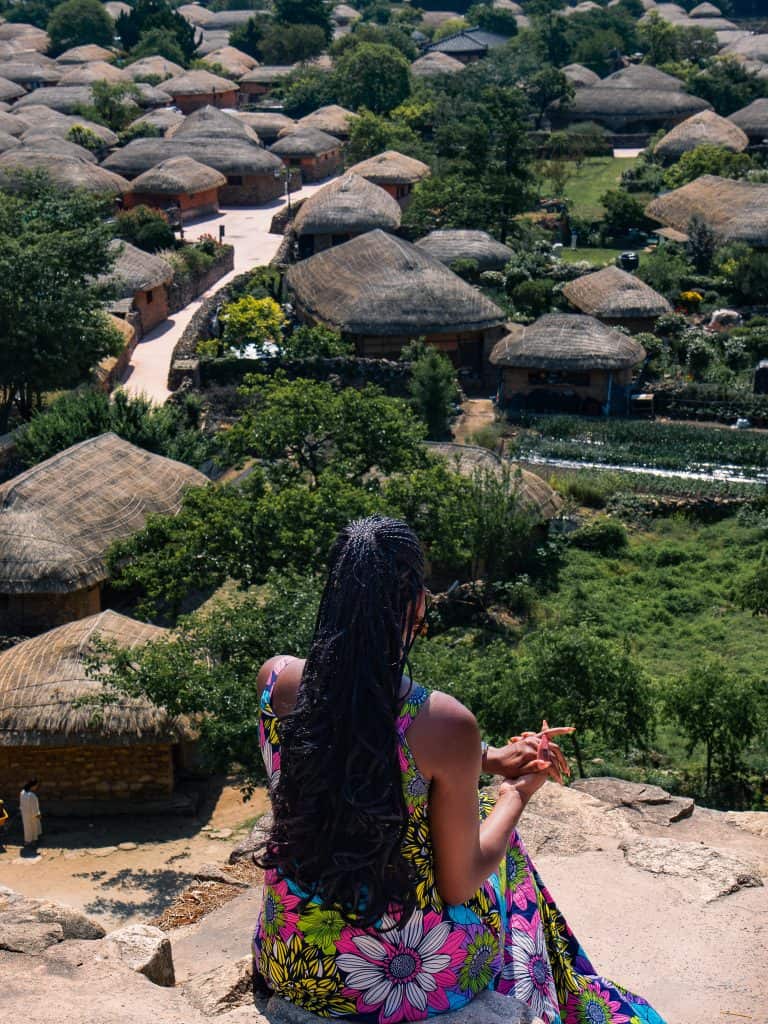 A woman with long black hair wearing a colorful dress sits on a rock overlooking a traditional Korean village with thatched-roof houses and lush greenery.