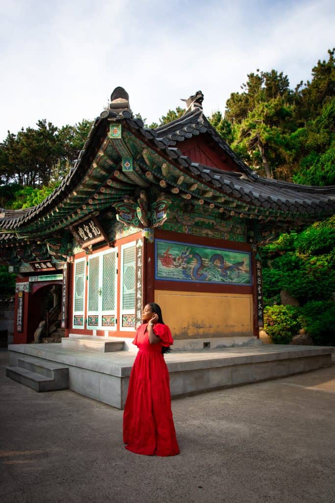 A woman in a red dress standing in front of a traditional Korean temple with ornate roof details and colorful artwork, surrounded by lush greenery.