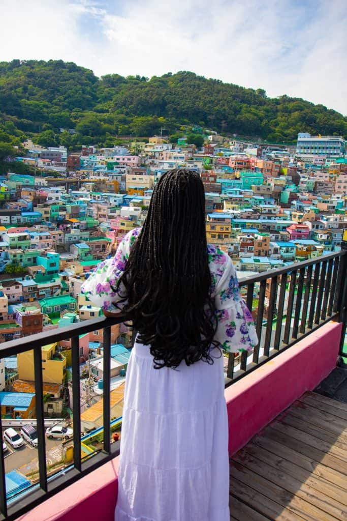 A woman with long black hair wearing a white dress and floral top overlooks the vibrant, colorful houses of Gamcheon Culture Village in Busan, South Korea from a balcony.