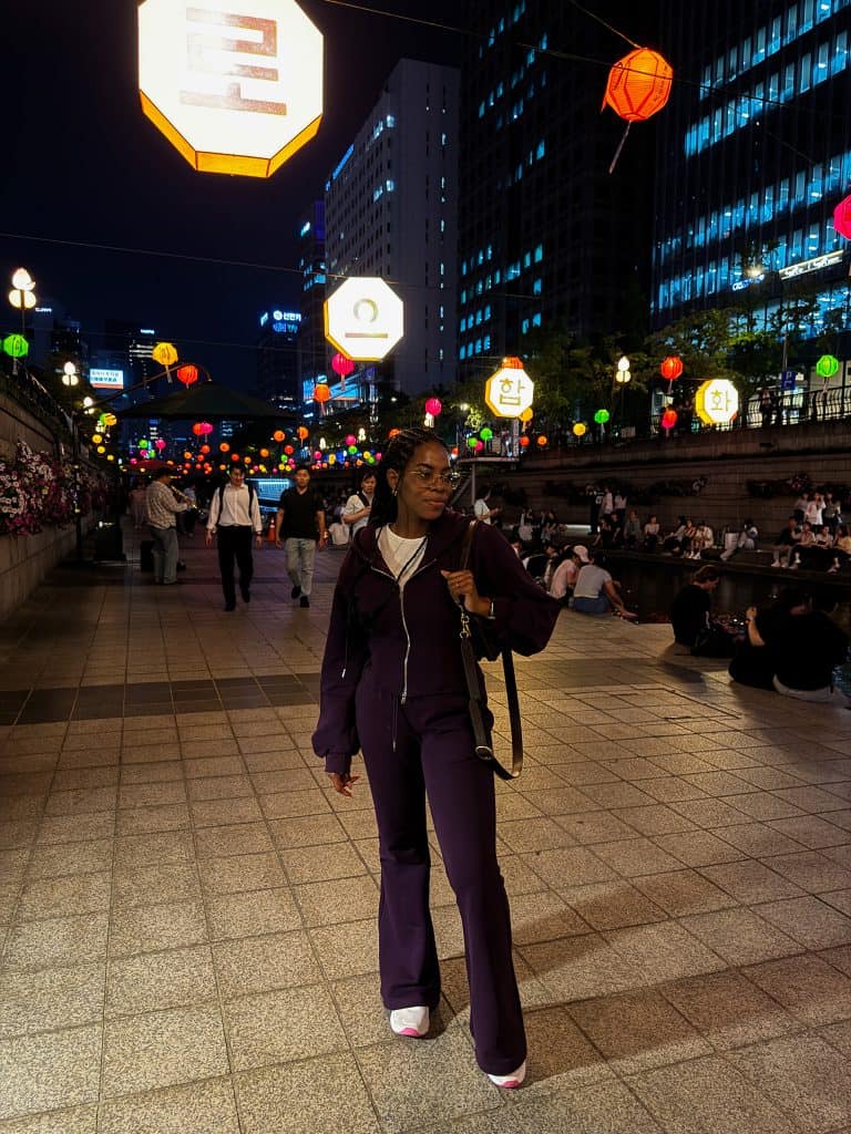 A woman walking through a vibrant night street in Seoul, illuminated by colorful lanterns and city lights, with people sitting and enjoying the lively atmosphere.