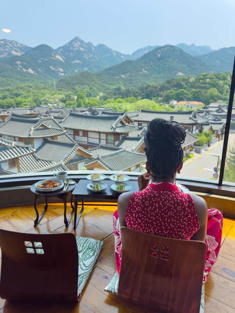 Nma Okafor sitting by a large window overlooking traditional Korean rooftops and mountain scenery in South Korea.