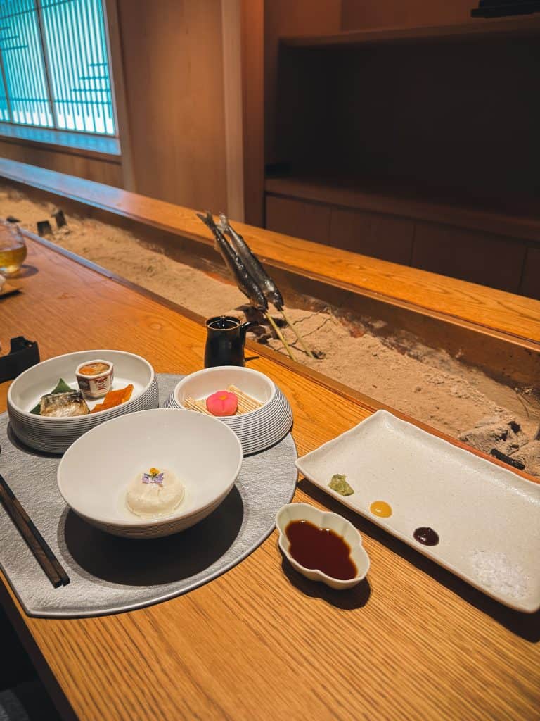 Elegant Japanese kaiseki meal served on a wooden counter with various small dishes, sauces, and a sand garden in the background.