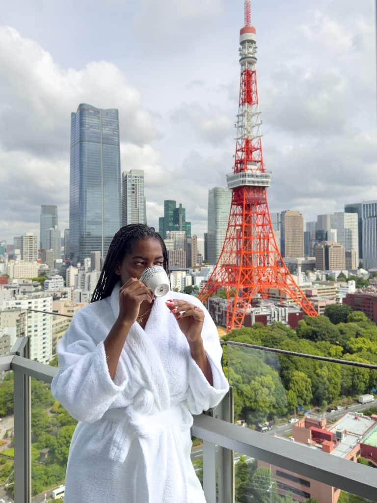 Nma Okafor wearing a white bathrobe and mask on a balcony with Tokyo Tower and cityscape in the background.