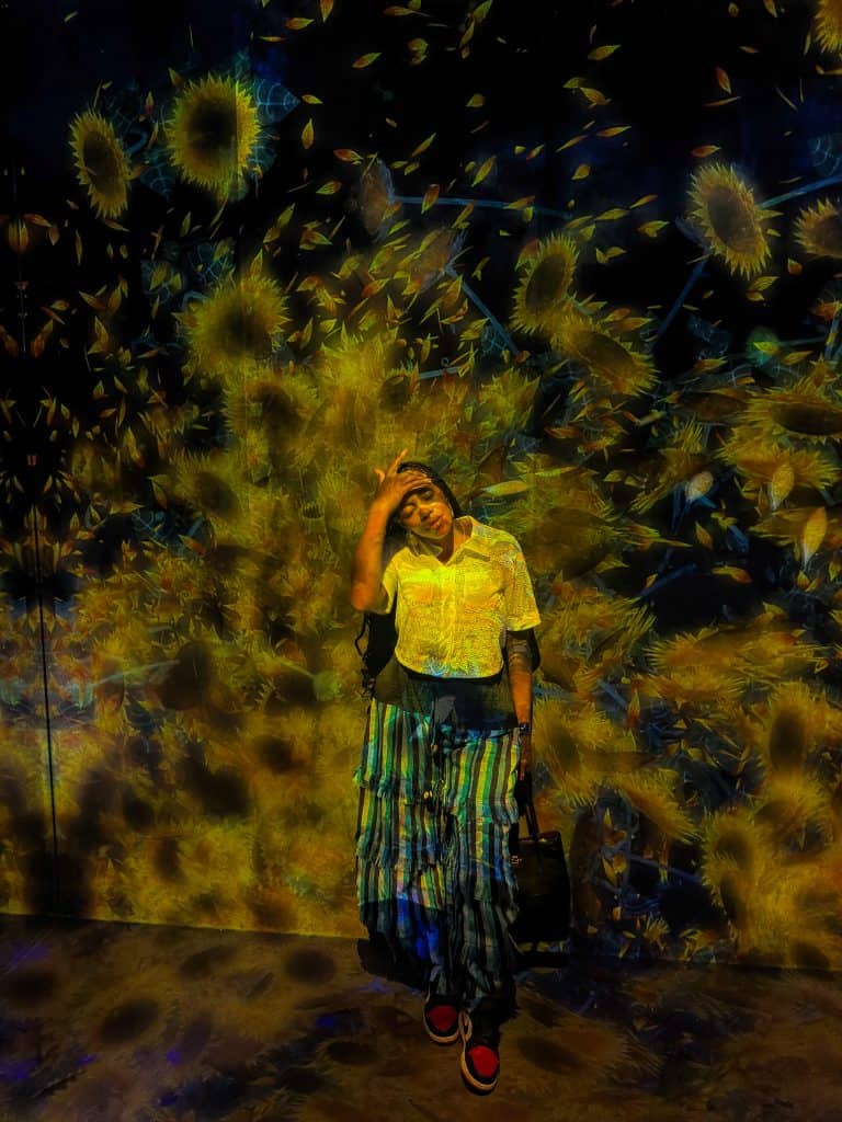 Nma Okafor standing in front of a vibrant sunflower-themed digital art display, wearing colorful clothing and sunglasses.